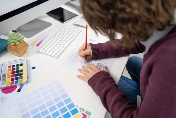Persona diseñando en papel frente a una computadora y paletas de colores